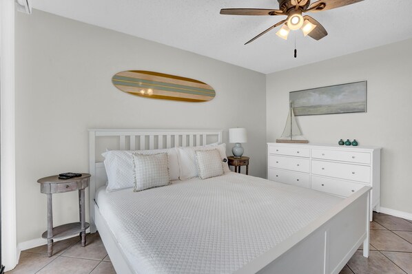 A bedroom with a large bed, a ceiling fan, a small side table, a dresser, and a wall-mounted painting. - Seaside Beach & Racquet 5917 (Orange Beach)