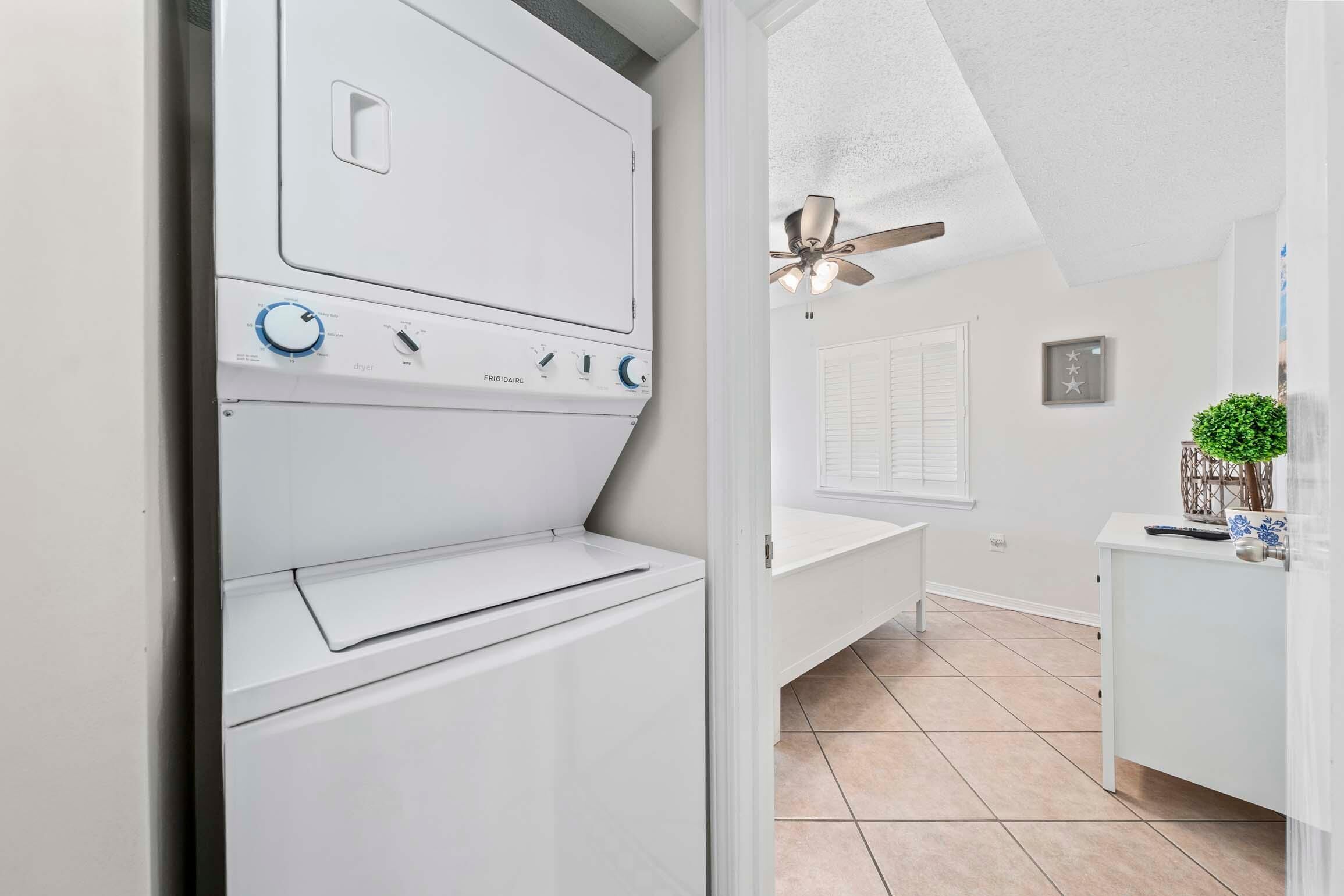 A laundry room with a front-loading washer and dryer, a ceiling fan, and a view into a bathroom with a sink and a potted plant.