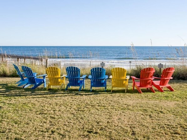 A row of colorful Adirondack chairs facing a beach with a wooden fence and sea grasses in the background. - Seaside Beach & Racquet 5917 (Orange Beach)