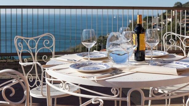 Ein Tisch für zwei Personen, bestückt mit weißen Metallstühlen, einer Weinflasche und leeren Weingläsern auf einem Balkon mit Meerblick.
