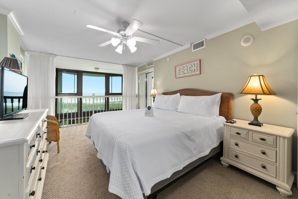 A bedroom with a large bed, a nightstand with a lamp, a dresser, a television, and a balcony with a view.