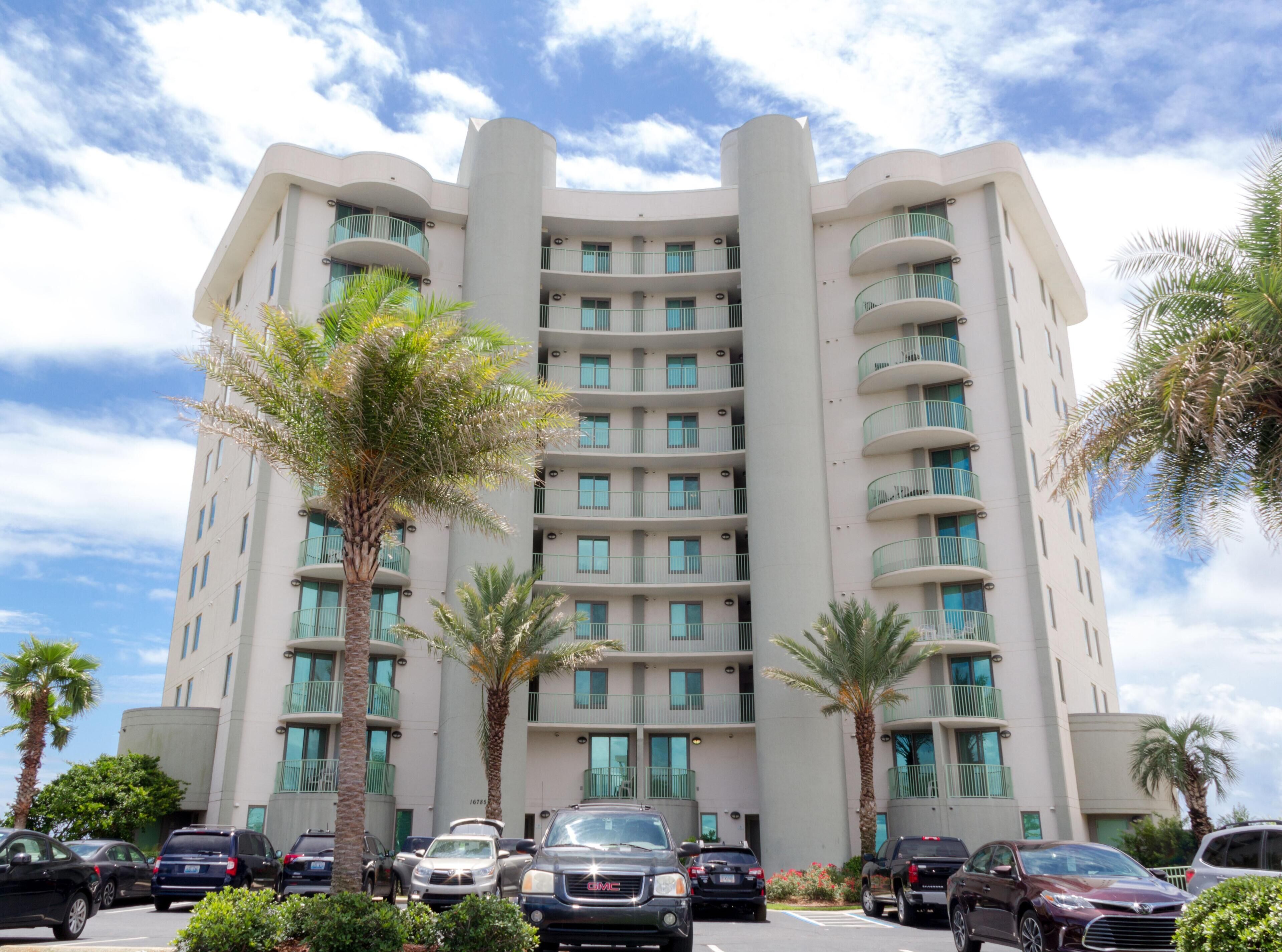 A multi-story hotel with a modern design, featuring balconies on each floor, a parking lot in front, and palm trees.
