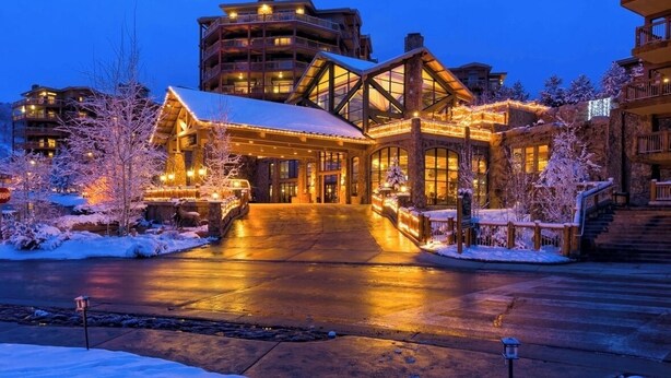Una cabaña con entrada cubierta de nieve, un área exterior techada y un edificio de varios pisos al fondo.