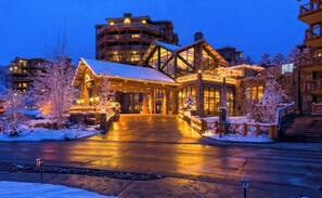 Un chalet dont l’entrée est recouverte de neige, doté d’un espace extérieur abrité et surmonté d’un bâtiment à plusieurs étages en arrière-plan.