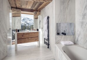 Ein modernes Badezimmer mit Holzboden, Marmorwänden, einem großen Spiegel, einer Badewanne und Blick auf schneebedeckte Berge.