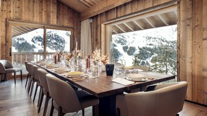 Un comedor con una larga mesa de madera dispuesta para seis personas, rodeada de sillas, y grandes ventanales que ofrecen vistas a un paisaje montañoso nevado.