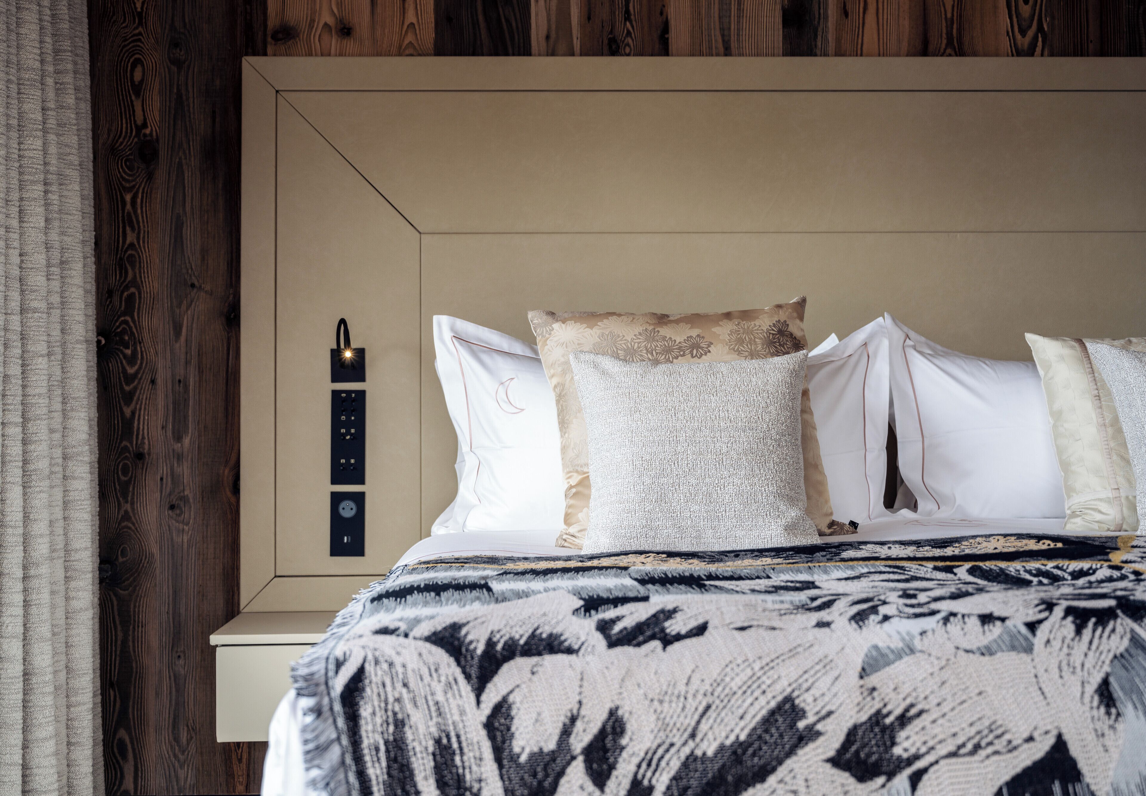 Een net opgemaakt bed met een gebloemd deken, een hoofdbord met bedieningspaneel en een houten wand.