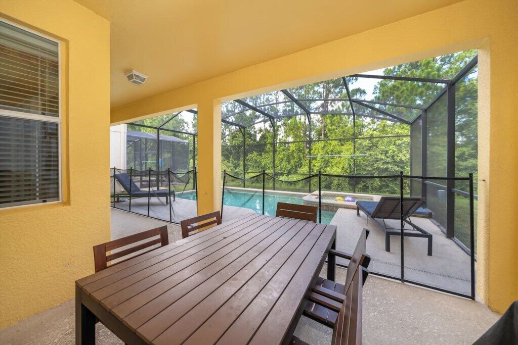 Een terras buiten met een houten tafel en stoelen, een zwembad en een glazen overkapping.