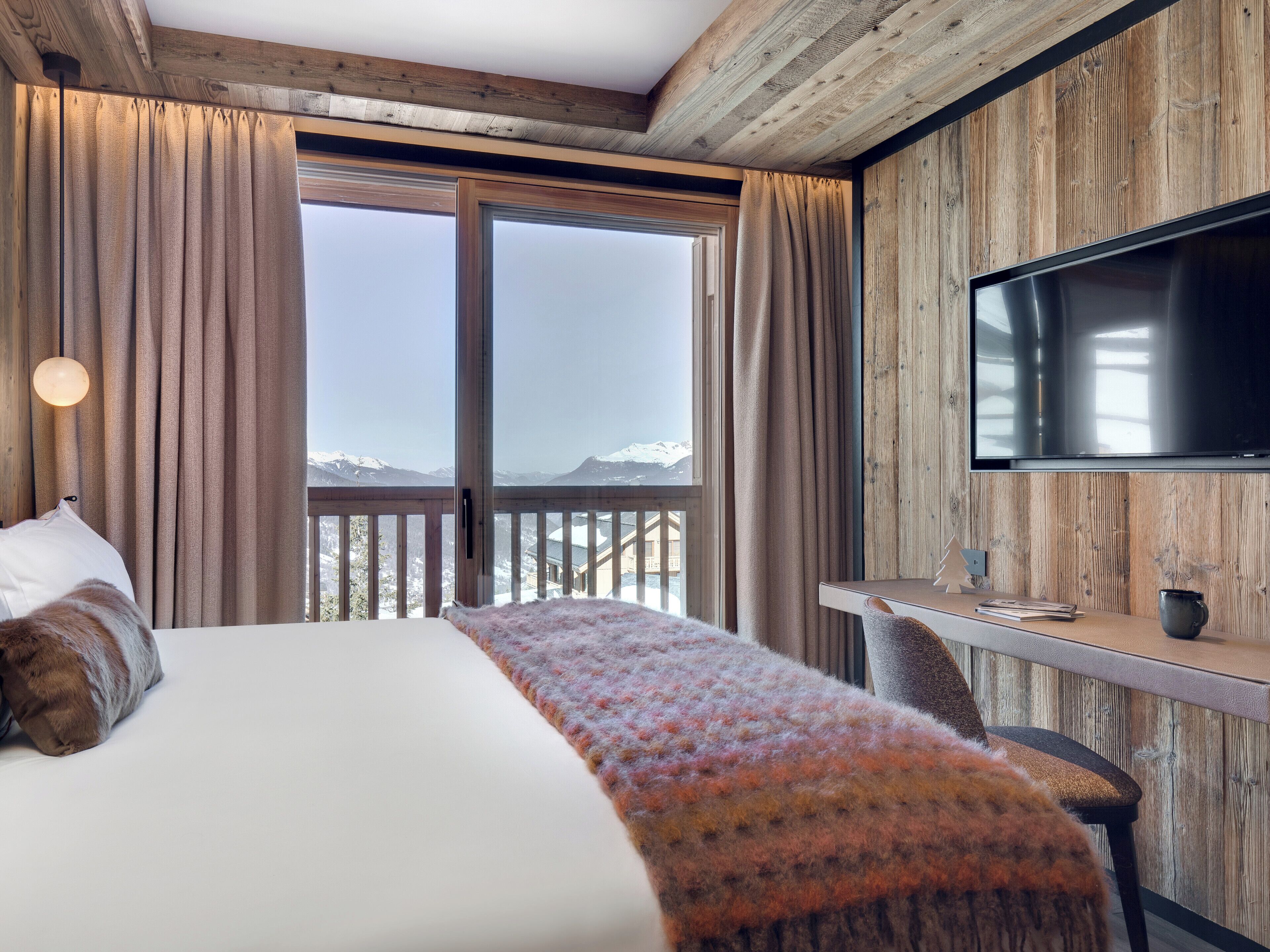 A bedroom with a large bed, a desk, and a TV. It features wooden walls and a view of mountains.