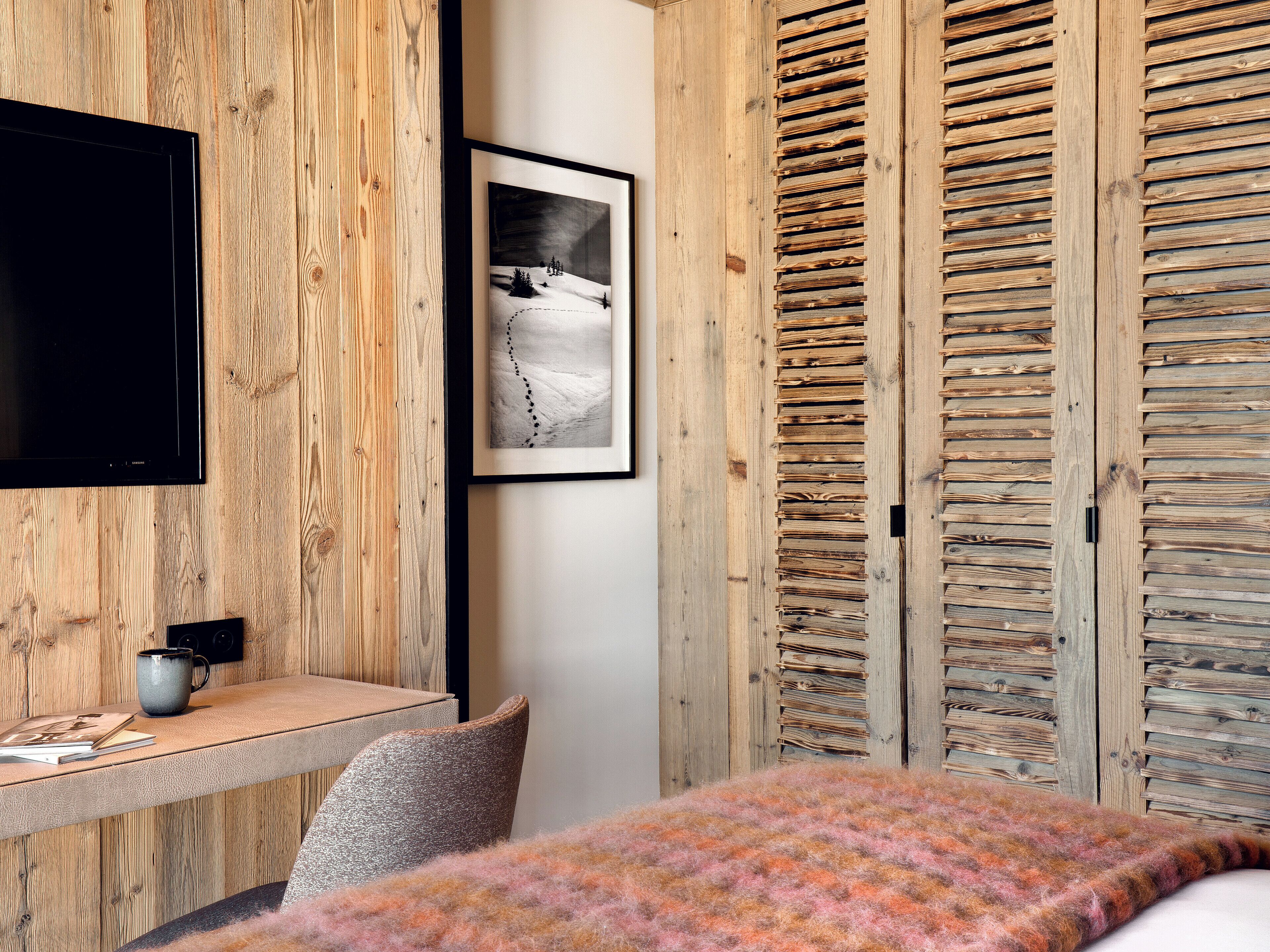 Een kamer met een houten wand, een wandtelevisie, een lijst met een afbeelding, een bureau met een mok en een bed met een gebreid plaid.