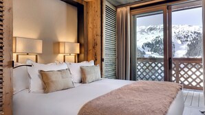 Un dormitorio con una cama grande, cabecera de madera, dos lámparas de noche y vistas a un paisaje montañoso nevado a través de una puerta corredera de vidrio.