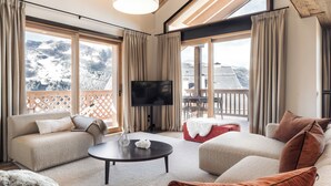 Ein geräumiges Wohnzimmer mit großem Fenster und Bergblick, einem Flachbildfernseher und einer gemütlichen Sitzecke mit einem roten Hocker.