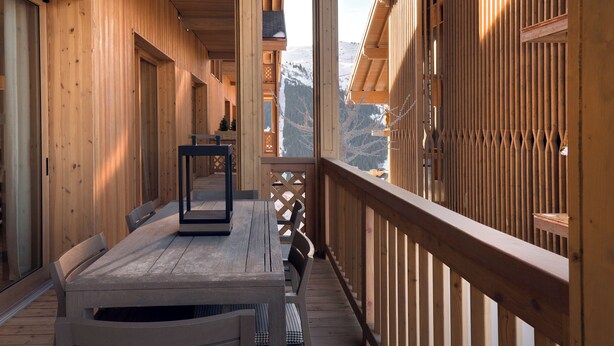 Un pasillo de madera con un banco y una mesa, que lleva a un balcón con vistas a montañas.