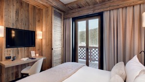 Habitación de hotel con paredes de paneles de madera, televisor de pantalla plana, escritorio con silla, cama con ropa blanca y balcón con vista a árboles.