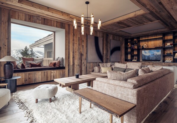 Ein gemütliches Wohnzimmer mit Holzvertäfelung, einem großen Fenster mit Blick auf eine verschneite Landschaft, einem modernen Sofa und einem Holztisch.
