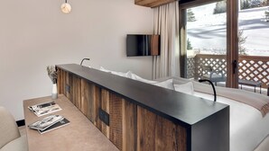 A modern hotel room with a wooden ceiling, a large window with a view of a snowy landscape, a flat-screen TV mounted on the wall, and a long wooden counter with magazines.