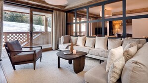 A cozy living room with a sofa, armchair, and coffee table, large windows with curtains, and a wooden ceiling.