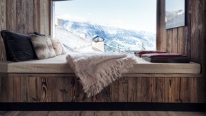 A cozy cabin interior with a wooden ceiling, a large window offering a snowy mountain view, a cushioned bench with pillows, and a framed picture on the wall.