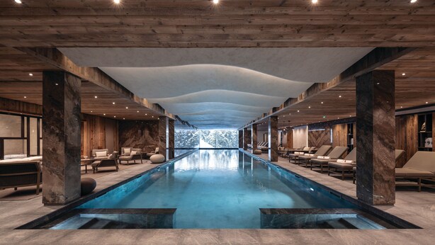 A modern indoor pool with a clear view of the outdoors, wooden ceiling beams, and lounge areas with seating.