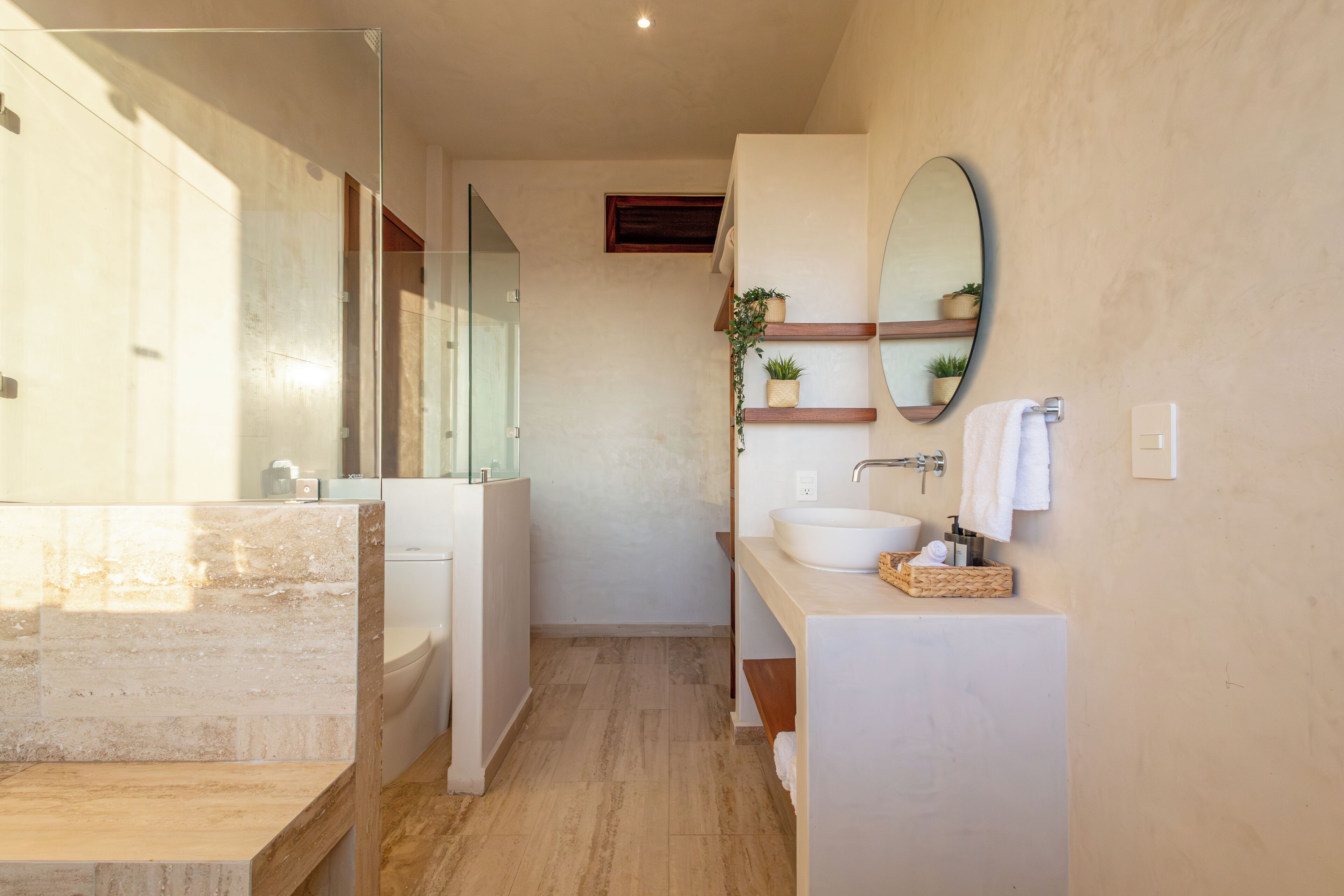 A modern bathroom with a glass shower enclosure, a large mirror, a sink with a wooden countertop, and a small potted plant.