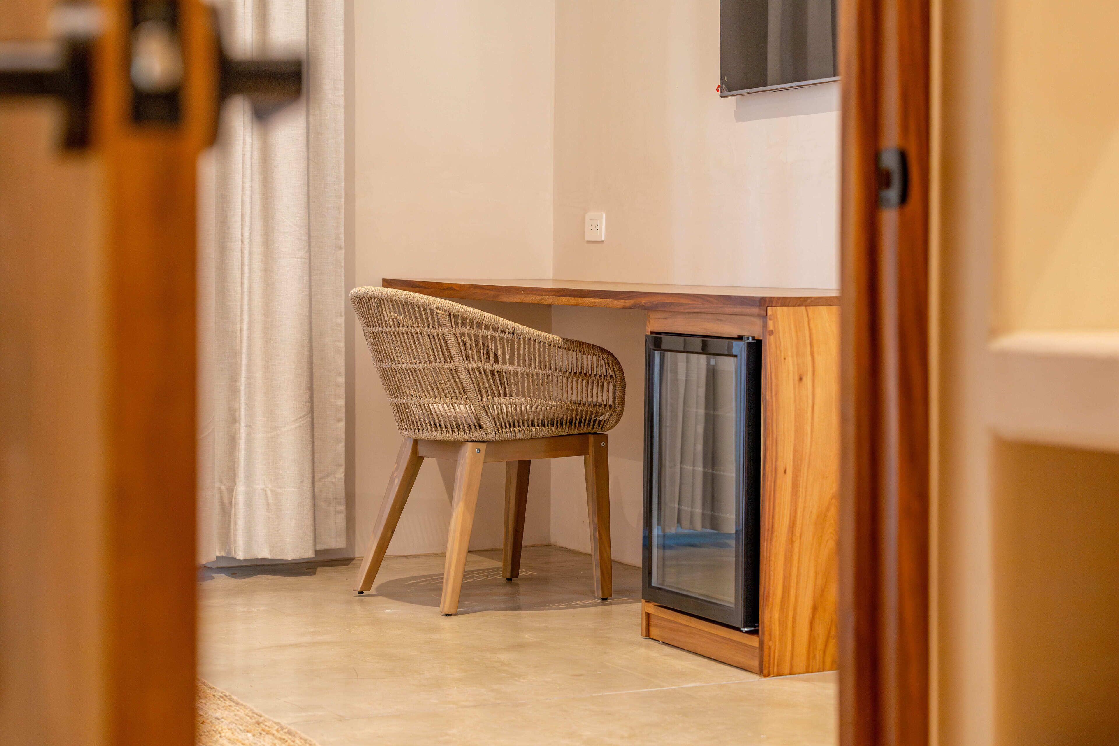 A room with a wooden desk, a wicker chair, and a built-in mini-fridge.