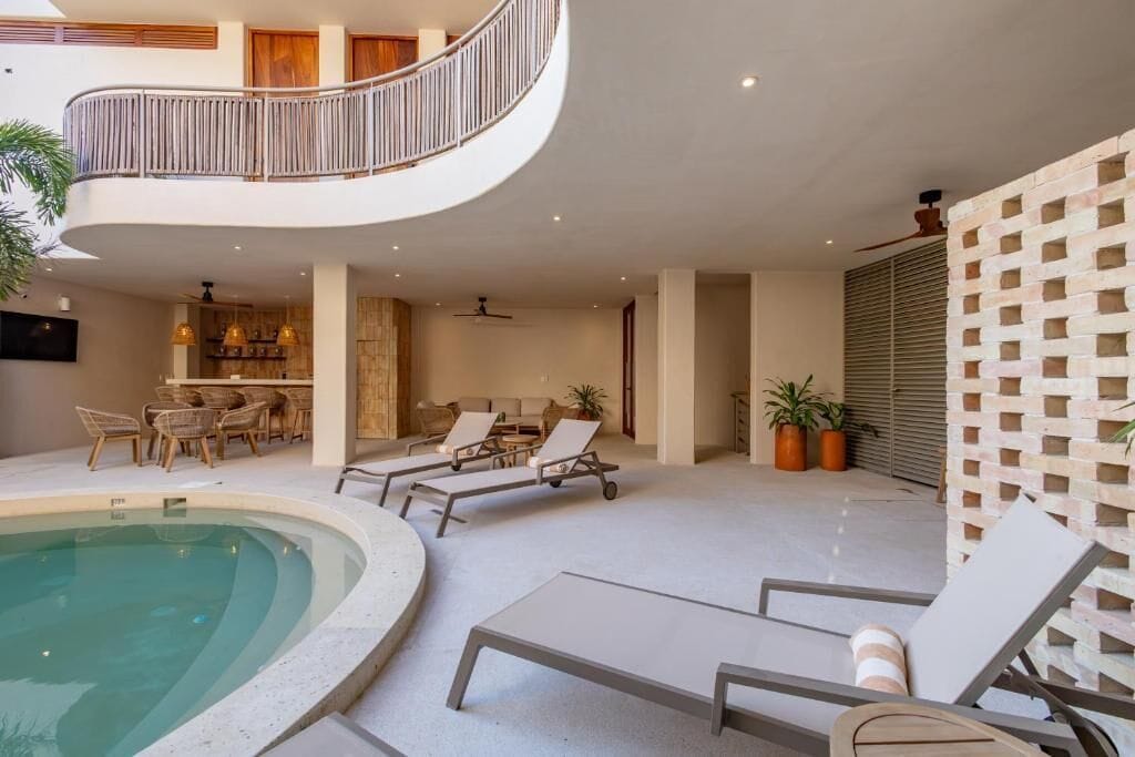 A modern indoor pool area with lounge chairs, a dining area, and a staircase leading to an upper level.