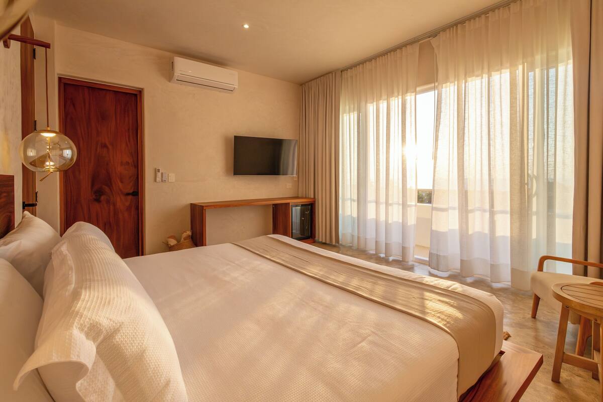 A hotel room with a large bed, a television, a wooden desk, and a window with sheer curtains.