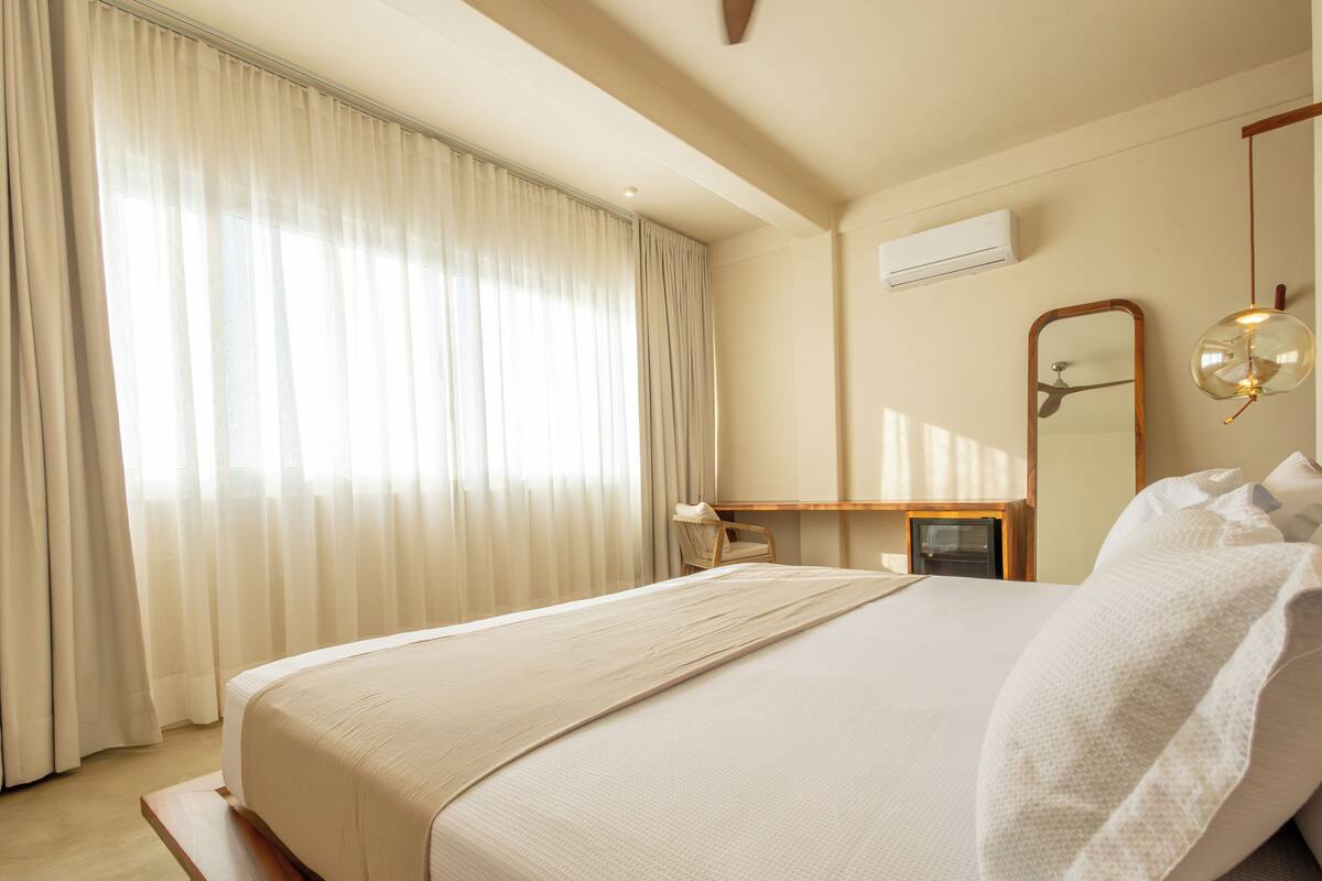 A hotel room with a large bed, a desk, a chair, a mirror, and a ceiling fan.