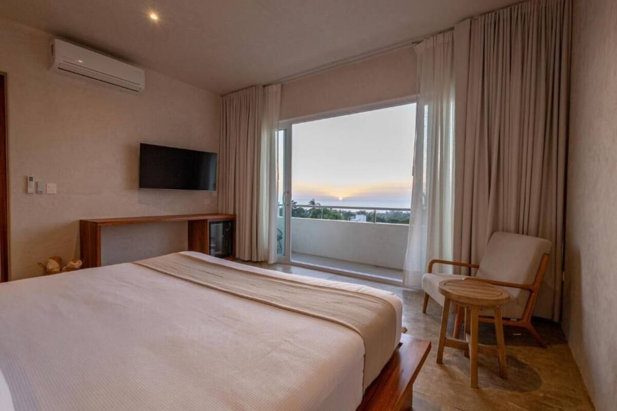 A hotel room with a large bed, a TV mounted on the wall, a balcony with curtains, and a chair.