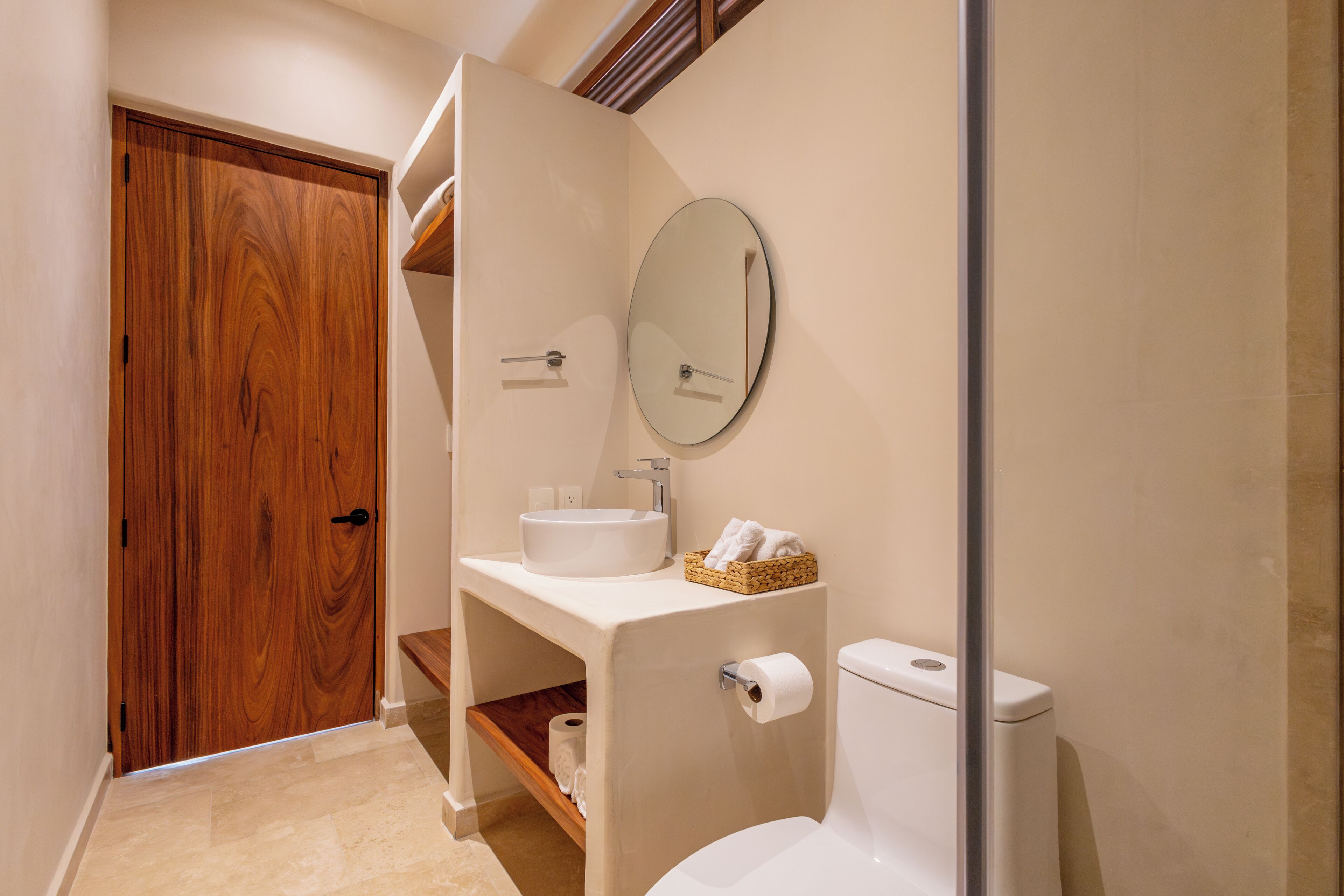 Un baño con una puerta de madera, un lavamanos blanco, un espejo redondo y un inodoro.