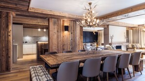 Un comedor espacioso con una gran mesa de madera y sillas modernas, una zona de estar acogedora con un banco, y una cocina al fondo.