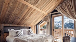 Un dormitorio con techo de madera, una cama grande con muchas almohadas, vistas a las montañas desde un balcón y una silla junto a la ventana.