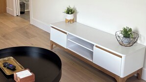 A modern living room with a flat-screen TV mounted on the wall, a white console table with two potted plants, and a black coffee table with a remote control and a small box.