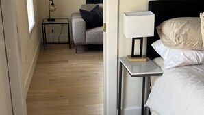 A hotel room with a bed, bedside table, lamp, and a view of a living area with a sofa and curtains.