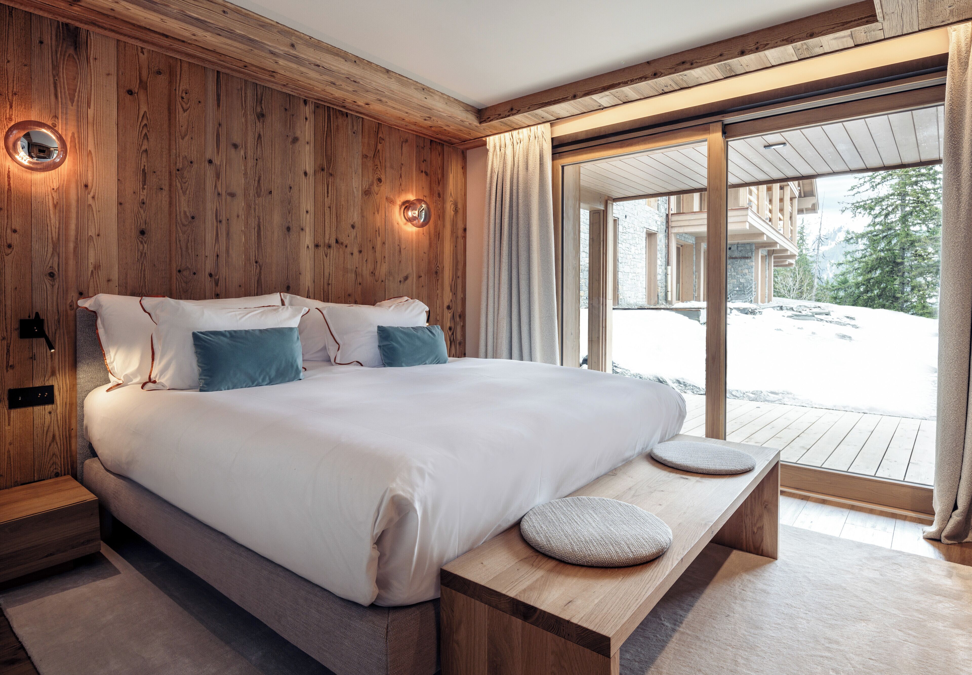 A modern bedroom with a wooden wall, a large bed with pillows, a bedside table, and a view of a snowy landscape through a glass door.