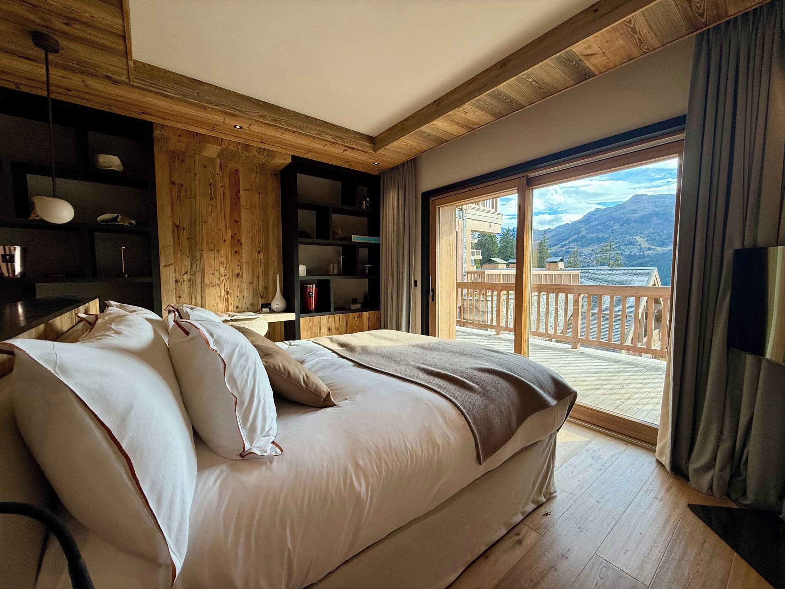 A bedroom with a large bed, wooden walls, a built-in shelf, and a balcony with a mountain view.
