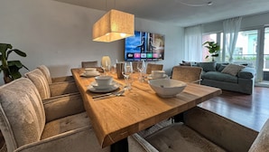 A modern dining area with a wooden table set for four, surrounded by cushioned chairs. A television is mounted on the wall, and a sofa is visible in the background. - Cozy, bright vacation apartment in trendy boho style #Cosiness Company (Cuxhaven)