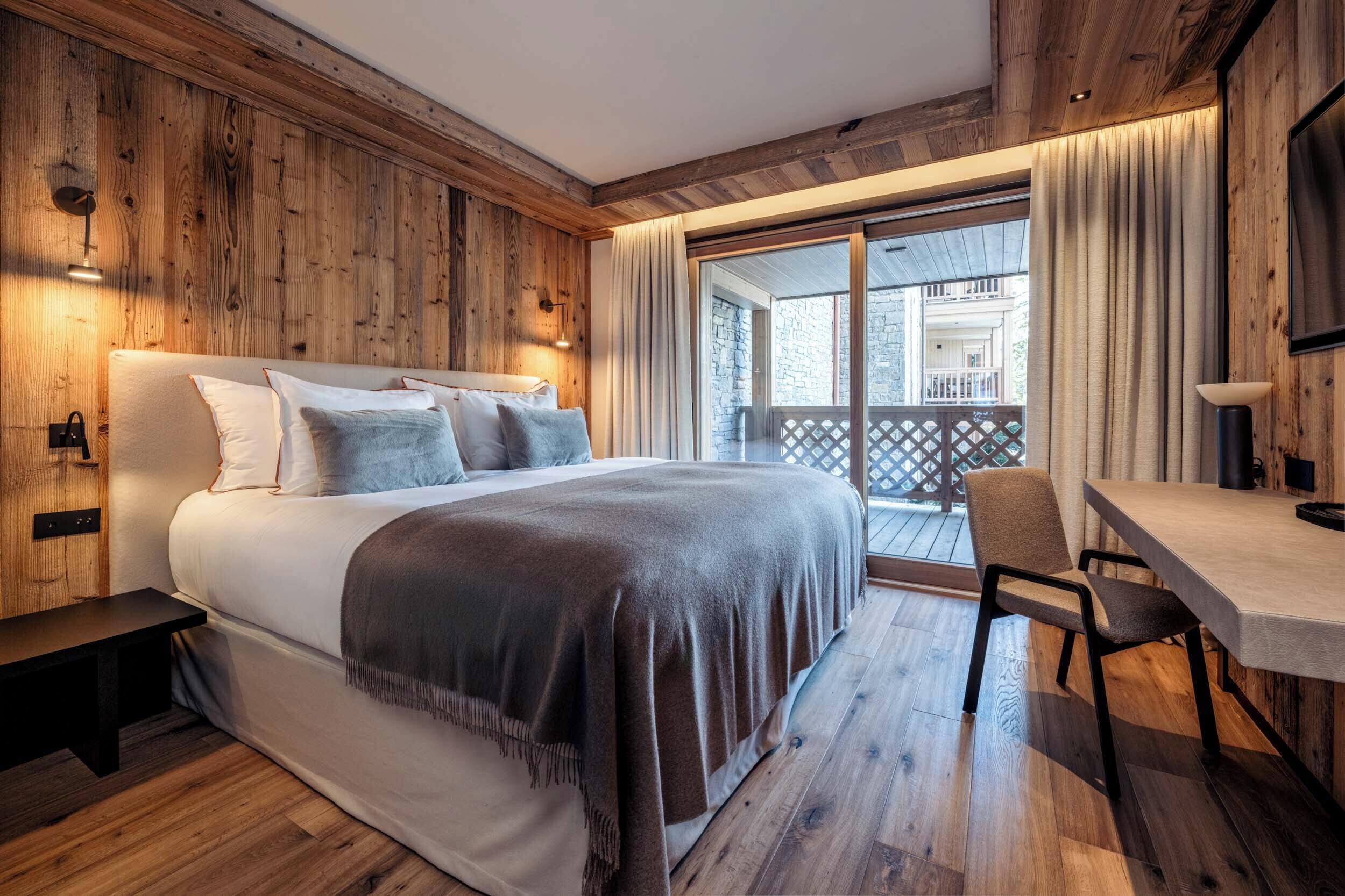Quarto com cama grande, paredes de madeira, varanda com vista e escrivaninha com cadeira.