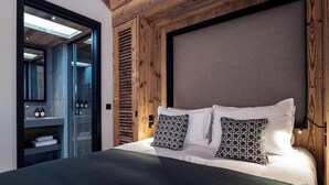 A modern bedroom with a wooden ceiling, a large bed with patterned pillows, a nightstand with bottles, and a glass shower enclosure.
