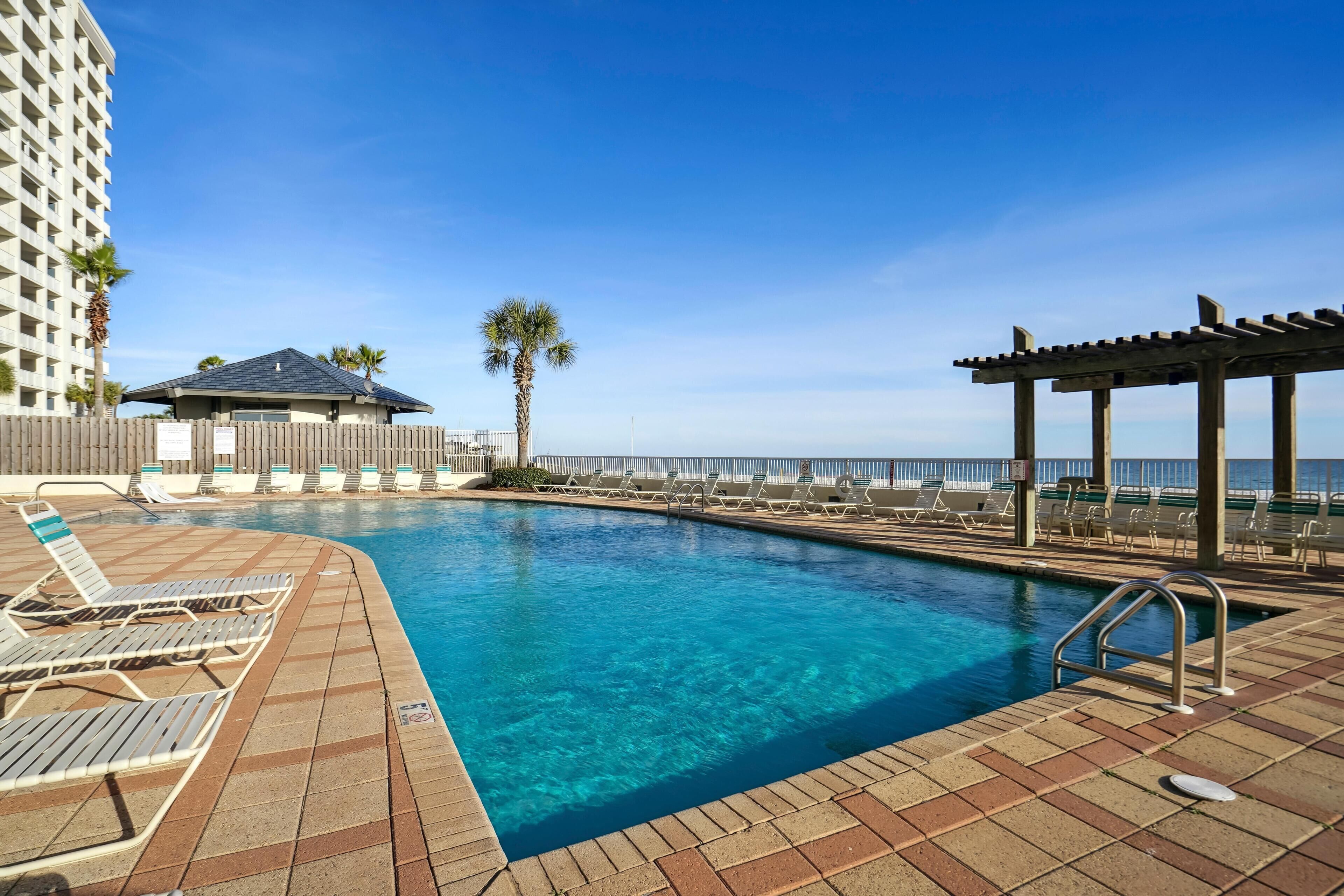 A swimming pool with lounge chairs and a pergola, surrounded by a high-rise building and palm trees.