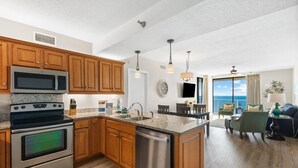 A modern kitchen with wooden cabinets, stainless steel appliances, and a large island. There is a dining area with a view of the ocean. - Shoalwater 1202 (Orange Beach)