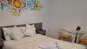 A neatly made bed with a white floral bedspread, two pillows, and a folded towel. There is a small bedside table with a remote control, a decorative wall piece, and a small round table with a chair.