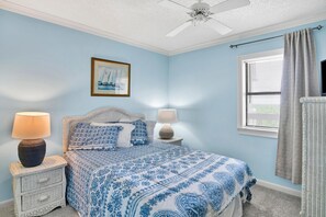 A bedroom with a bed, two nightstands, a ceiling fan, a window with blinds, and a framed picture on the wall. - Shoalwater 903 (Orange Beach)