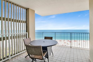 A balcony with a round table and two chairs, overlooking a beach with umbrellas and people. - Shoalwater 903 (Orange Beach)