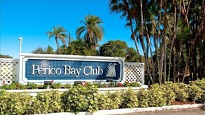 A sign for the Perico Bay Club with a clear view of the surrounding palm trees and a white fence.