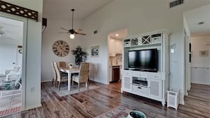 A spacious living room with a wooden floor, a dining area with a round table and chairs, a ceiling fan, and a TV on a white entertainment unit.