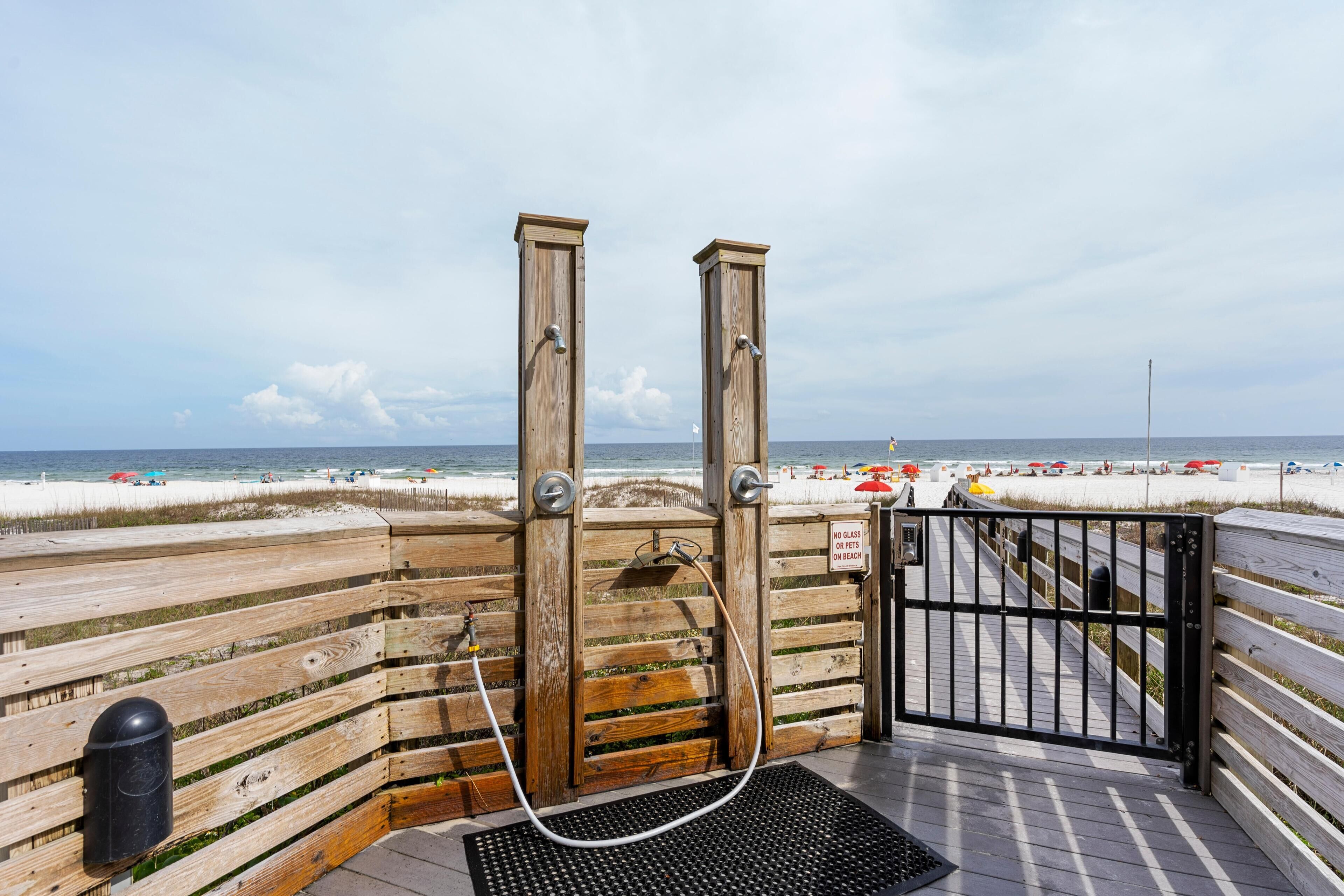 A wooden boardwalk leading to a beach with a wooden structure, a black gate, and a sign prohibiting glass containers on the beach.