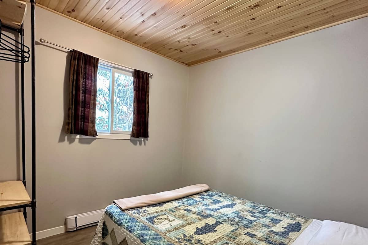 A room with a bed, a window with curtains, a radiator, and a wooden ceiling.