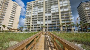 A multi-story hotel with a central atrium, balconies, and a wooden boardwalk leading to the entrance. - White Caps 1502 (Orange Beach)