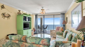 A living room with a green cabinet, a sofa, a glass coffee table, and a view of the ocean through large windows. - White Caps 1502 (Orange Beach)